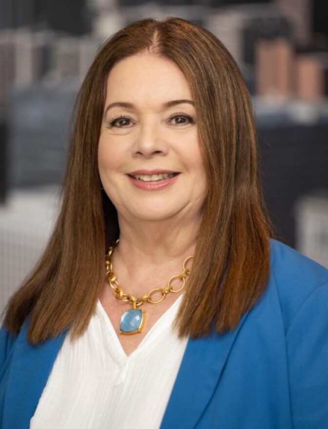 a woman with brownish-red hair wearing a blue suit jacket with a white blouse and a gold necklace with a blue jewel