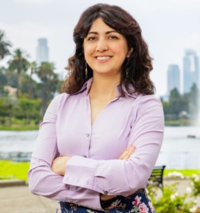 a woman with shoulder-length brown hair wearing a purple-pink blouse with a park behind her