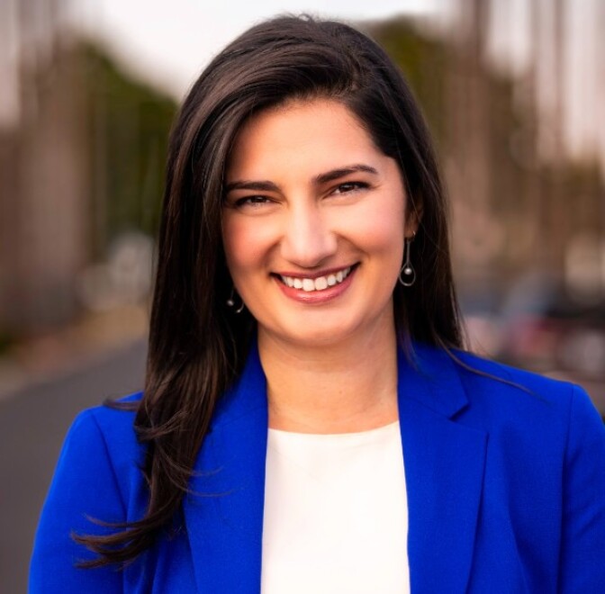 a woman with long brown hair wears a blue blazer over a white blouse with earrings