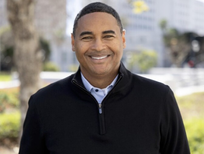 a man with short dark hair wearing a black pullover with a white collared shirt underneath