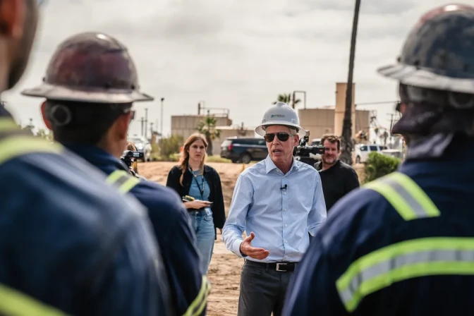 A man with light skin tone, wearing a safety helmet and shades, speaks to workers wearing safety helmets and vest in the foreground.