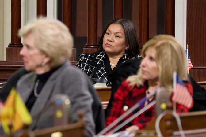 Assemblymember Leticia Castillo, a woman with medium skin tone, wearing a checkered coat, sits and listens to someone out of frame. Two woman sitting in front of Castillo are out of focus looking in the same direction..