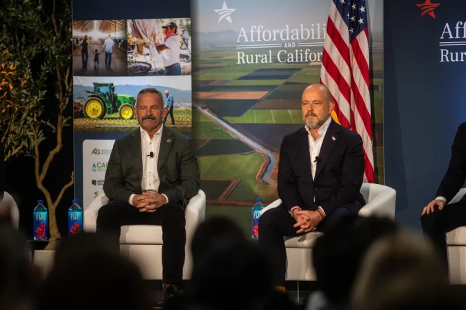 Chad Bianco, a man with light skin tone, wearing a gray suit jacket and white shirt, sits on a chair next to Steve Hilton, a man with light skin tone, wearing a black suit and white shirt, on a stage with a crowd of people listening in the audience, who are out of focus in the foreground. Singage behind them shows photos of farmers and text that reads "Affordability and rural California."
