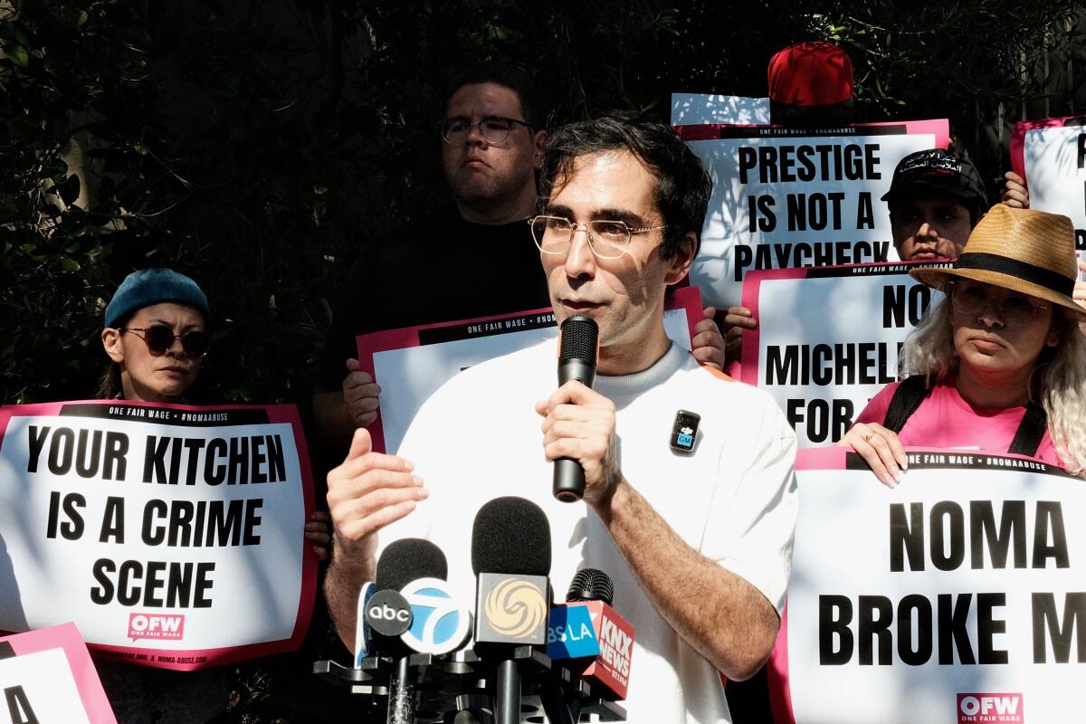 A man at a microphone stand addresses the press. He is flanked by fellow protesters holding signs