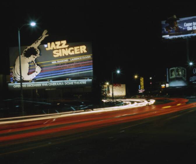 A color photograph of a street scene from 1980 at night. Billboards line the street, including one advertising for Jazz Singer and one for Marlboro cigarettes.