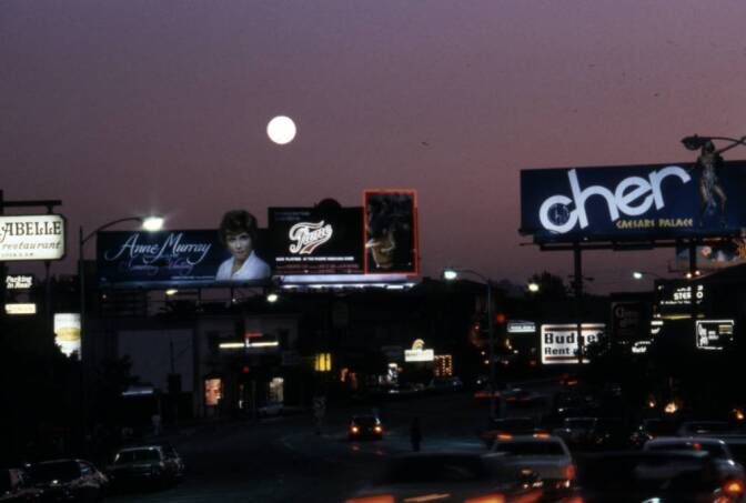 A night scene on a busy street. The moon is full. And cars are packed on the street. A number of billboards line the street.
