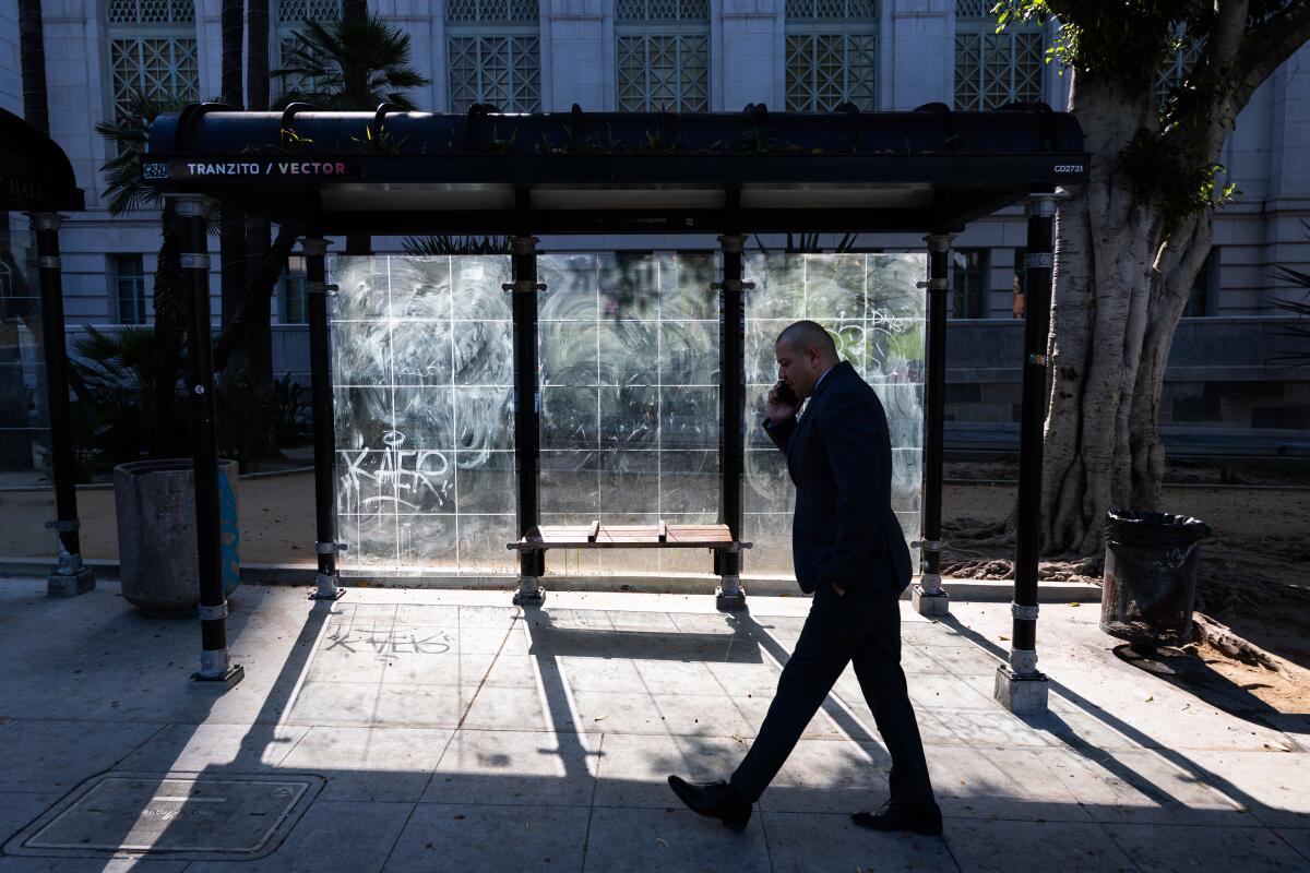 A pedestrian walks by a bus stop, marked by graffiti and dirty windows in front of Los Angeles City Hall.