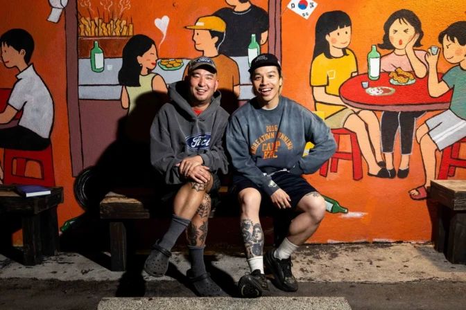 Two Asian men, both wearing hats and active gear, sit on a bench smiling and posing for a photo in front of a mural.