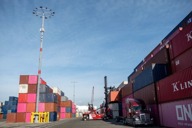 Stacks of shipping containers of various colors are seen under blue skies with a crane in the background.