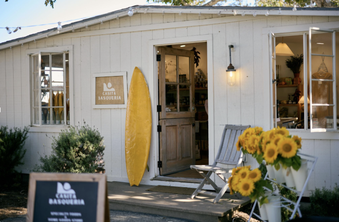 The exterior of a quaint cottage like building, with cream wooden paneling on the outside, a wooden front door, and a yellow surfboard leaning next to it, with yellow sunflowers in a jar in the front.