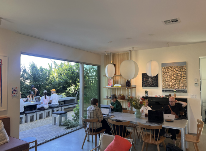 The interior of a living room has a silver dining table with people sitting around it; the atmosphere is mellow and stylish; outside the open patio doors are people sitting at another large table