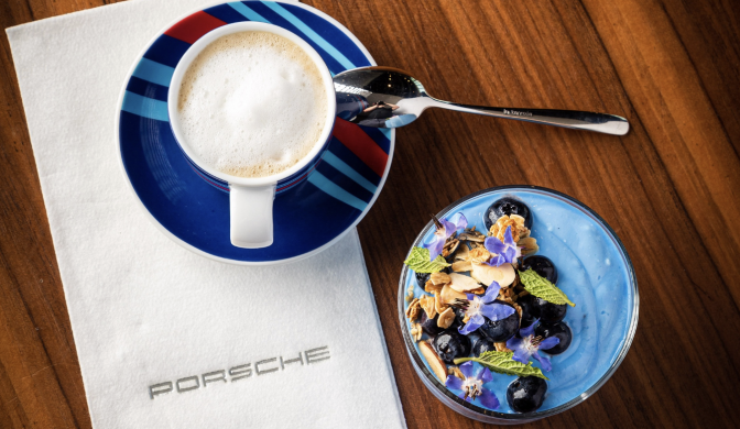 A blue and red cup of coffee with a foam top sits on top of a white napkin which says Porsche. To its side is a blue ceramic plate with a mix of blueberries, almonds and green mint leaves.