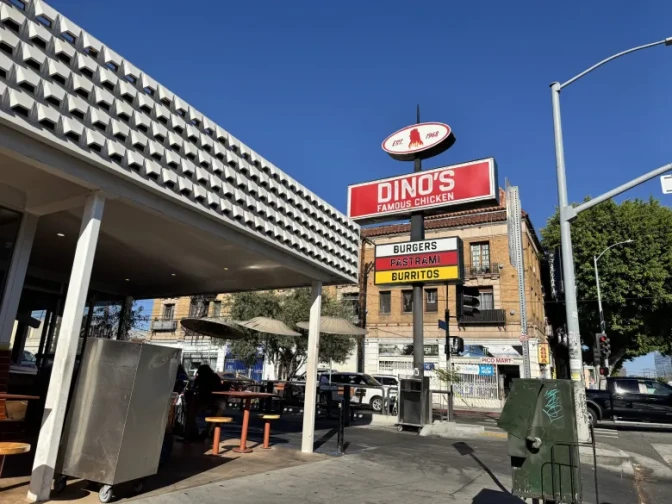 Signage that reads "Dino's Famous Chicken. Burgers. Pastrami. Burritos" stands on the corner next to a small building with outdoor seating and a street intersection.
