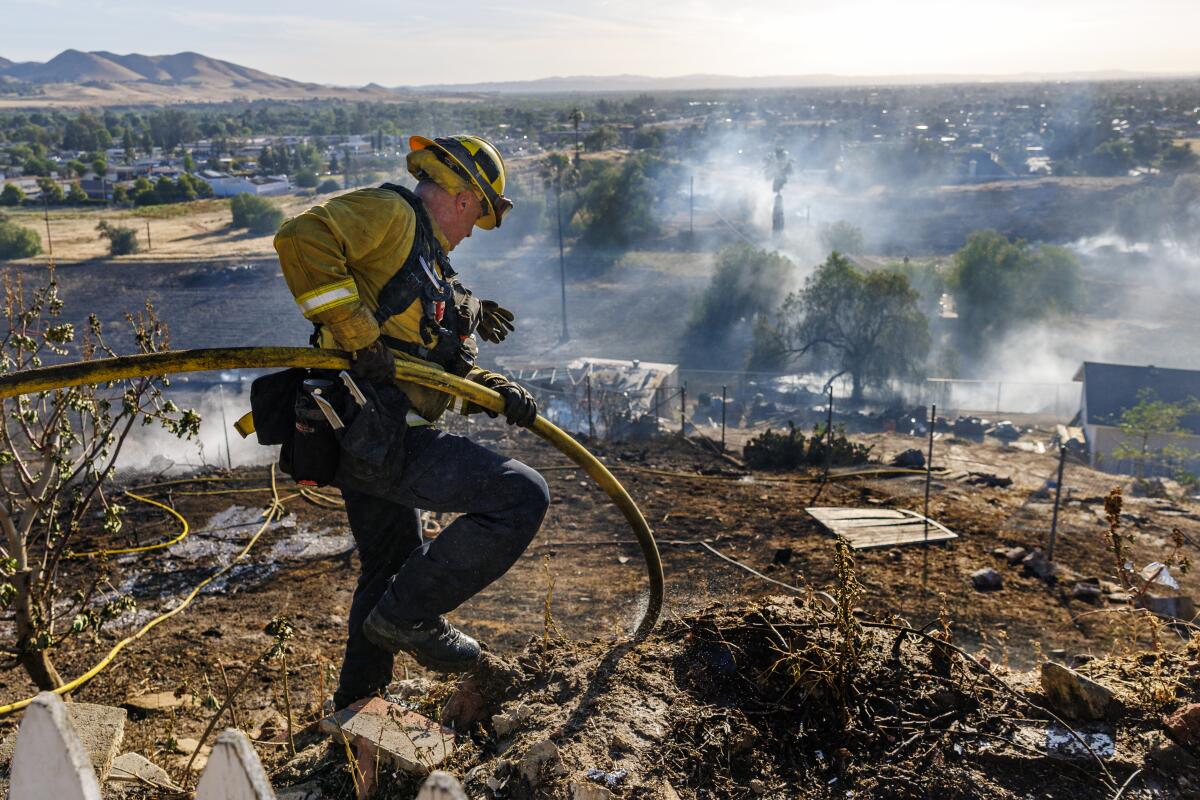 A Riverside County firefighter maneuvers his hose on a burned hillside behind a damaged home 