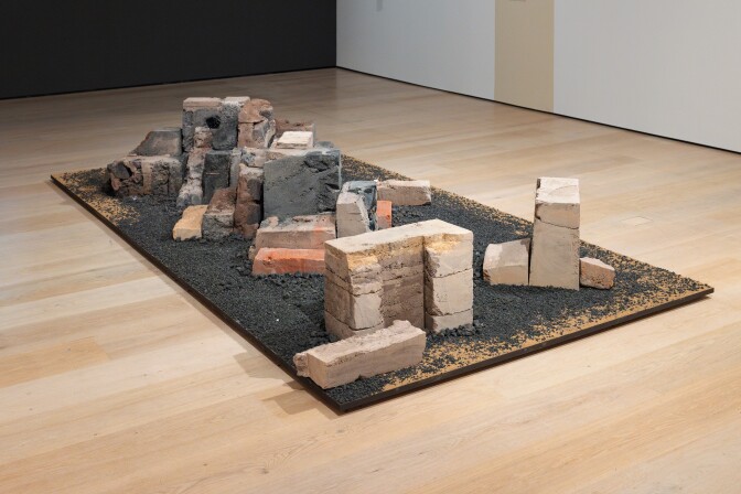 Stacks of red, brown and grey bricks in what appear to be random piles on a large plank of wood covered in a black gravel-like substance.