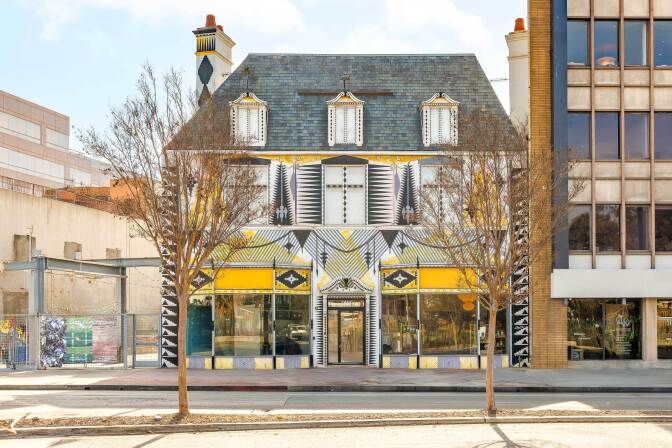 A street view of a three-story house-like building painted with white, black and yellow shapes and office buildings on either side of it. 
