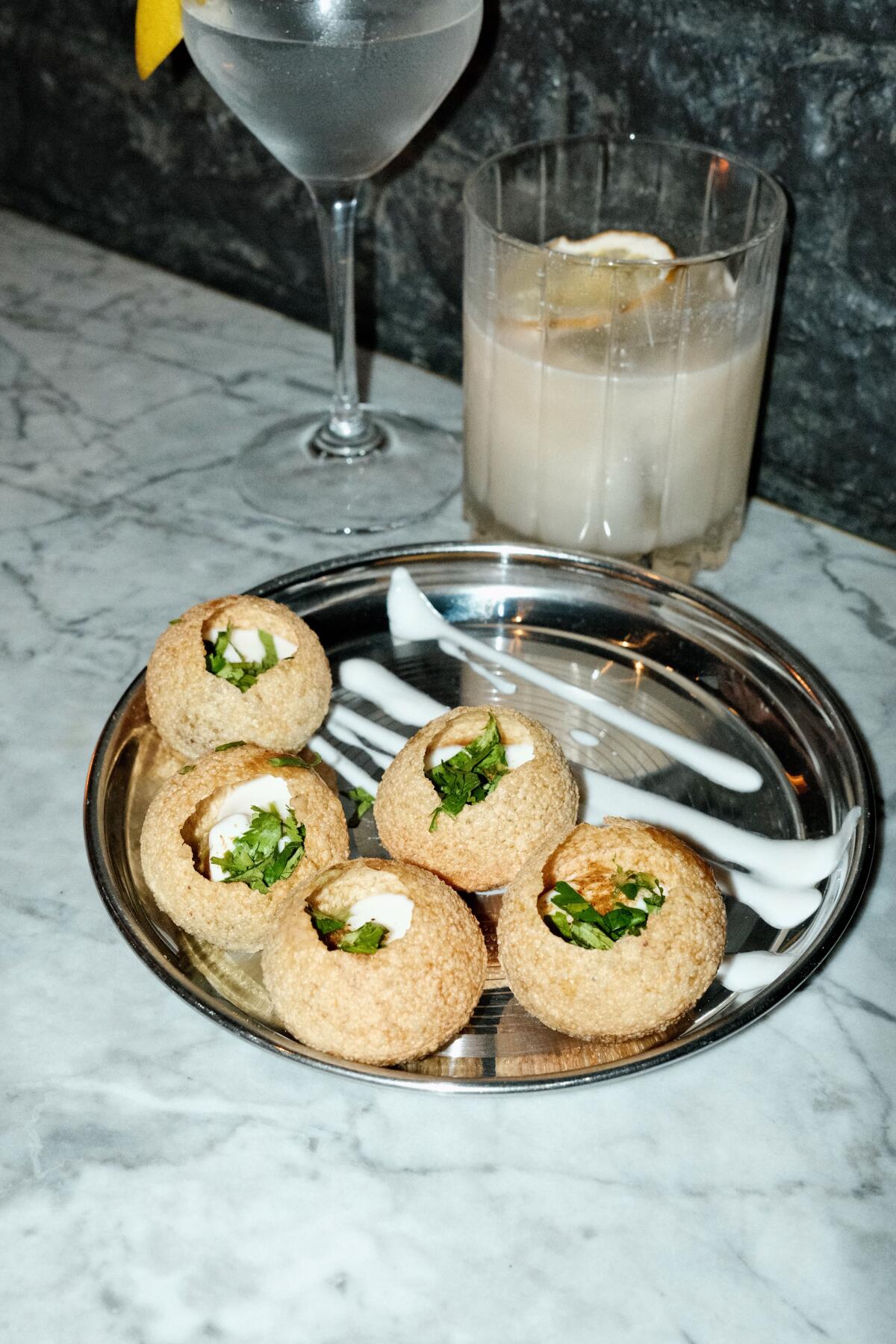 Dahi Poori, a chaat of hollow crackers, yogurt, tamarind and mint chutney, next to two cocktails on a marble table