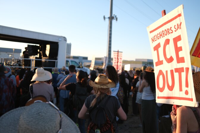 A crowd of people gather outside a building. One person hold a signs that states "ICE OUT!"