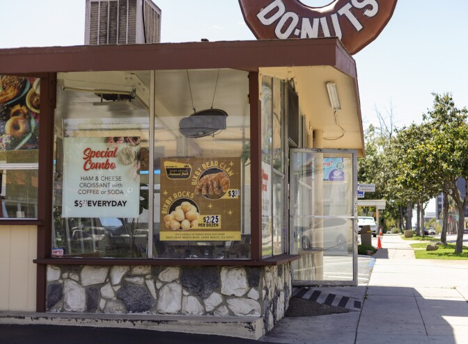 A wide look at the corner of the donut shop of the brown and stone building. It has signs in the window that advertise specials, including the Bixby Bear Claw and Donut Rocks. A giant brown donut is fixed to the roof.