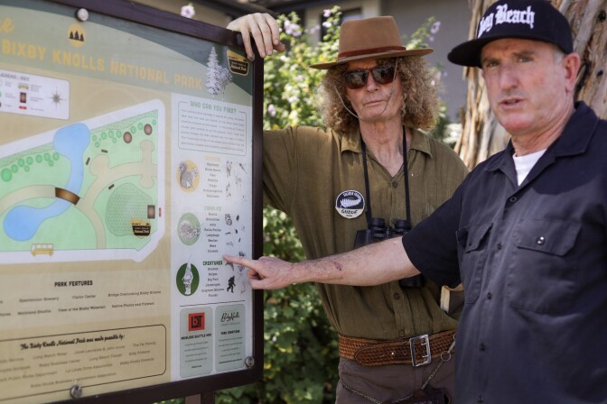 Two white men stand in front of a park sign that has a map on it and guide. The man on the left is dressed as a park ranger with a brown hat, sunglasses and binoculars on. The man on the right is wearing a black button up shirt and black hat. He's pointing to the sign while looking at the camera.