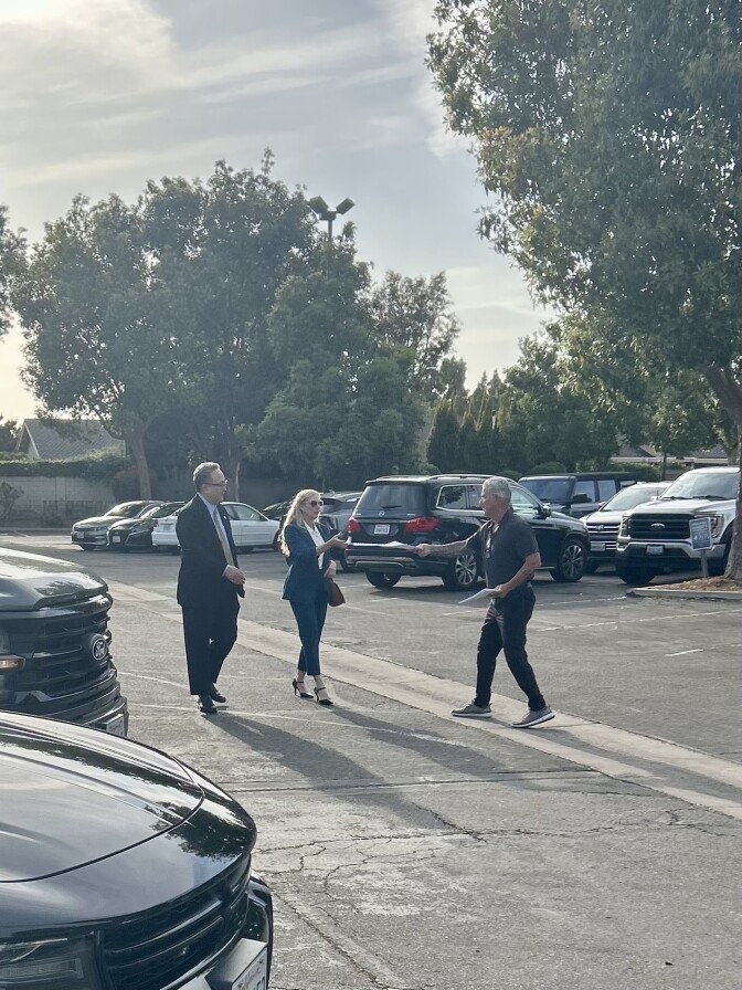 In a parking lot, a man hands a document to a woman in a suit who is walking with another man in a suit.
