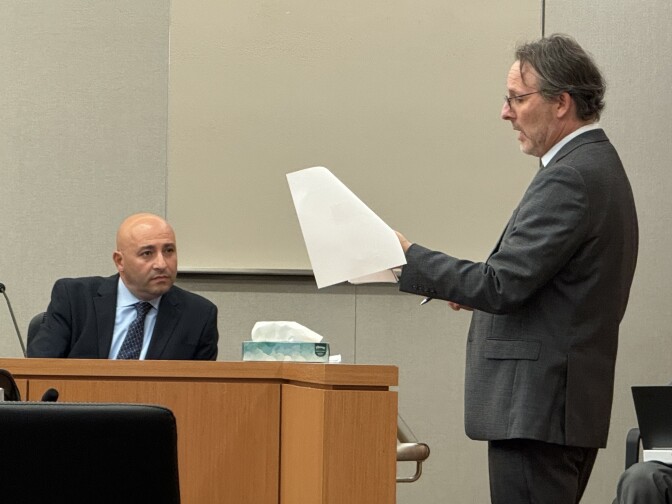 The setting is a courtroom: A man wearing a dark suit is sitting and looking at a man, also wearing a dark suit, as the man is speaking in reference to some papers in his hand.