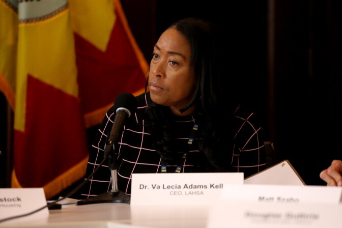 A Black woman sits at a dais with a flag in the background. A name placard in front of her reads: Dr. Va Lecia Adams Kell[um].