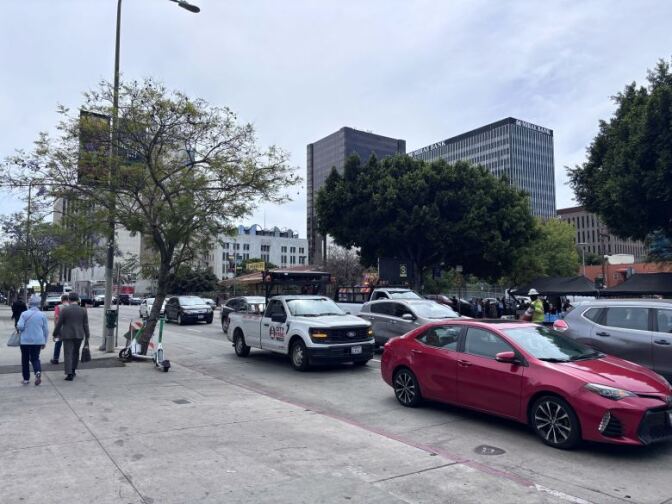 Cars wait in traffic on a street as people walk along a sidewalk. There are large buildings and trees in the distance.