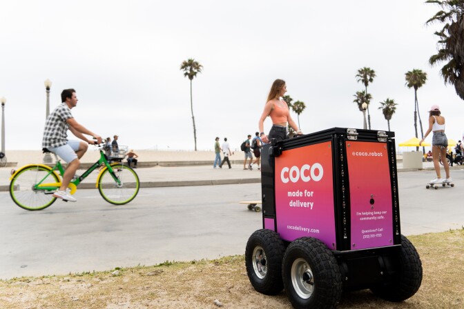 a black, box-shaped robot with four wheels and a pink and purple sign on the side that reads, "coco, made for delivery," sits outside a restaurant.