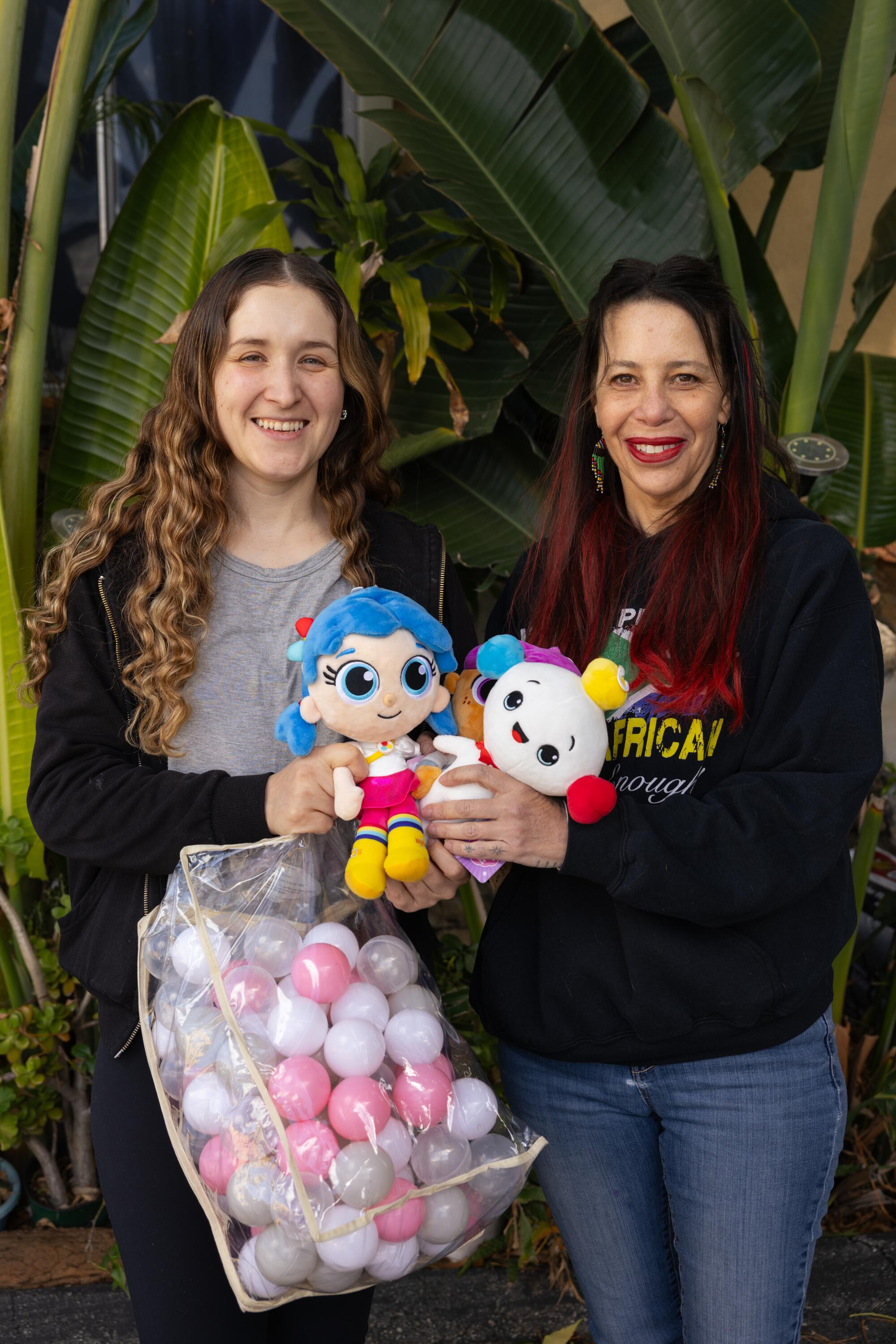 Two women stand in front of a green plant holding stuffed dolls and a bag of ball pit balls.
