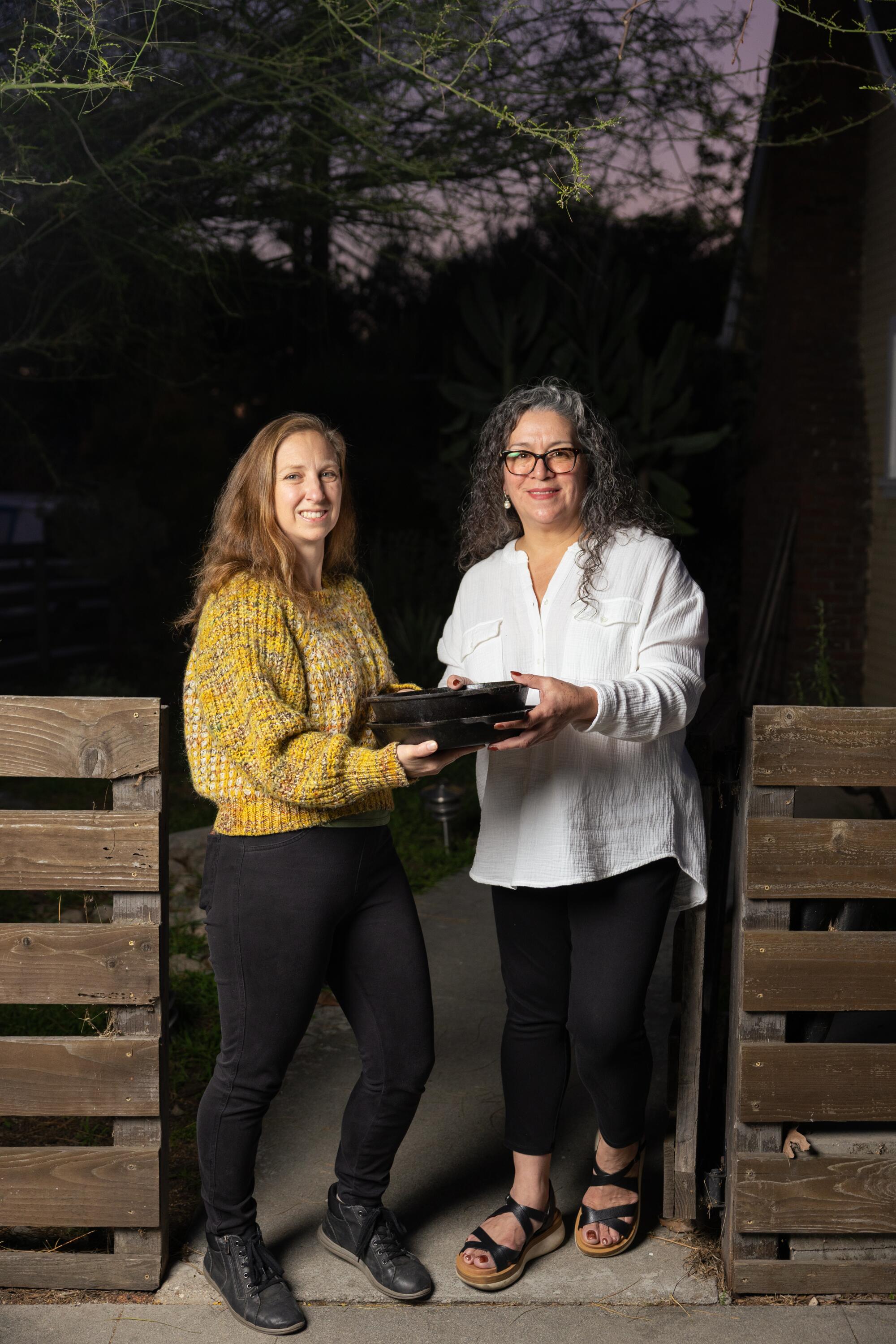 Two women stand by a gate at night holding cast iron pans.