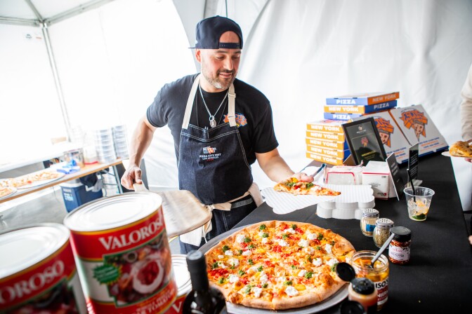 A man with a medium dark skin tone serves a slice at his booth inside the Pizza City Fest tent, with a large topped pizza on the table and stacked New York-style pizza boxes visible behind him.
