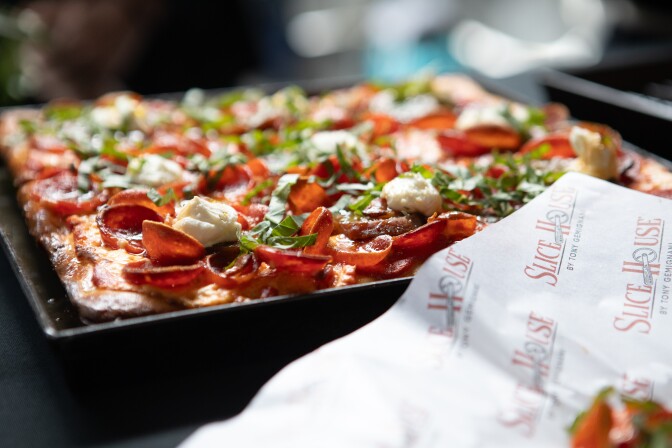 A rectangular Detroit-style pizza topped with cupped pepperoni, dollops of ricotta, and fresh basil, served in a black pan with a Slice House by Tony Gemignani wrapper in the foreground.
