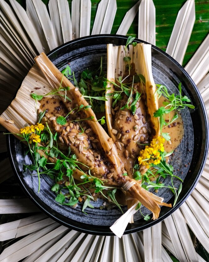 Two tamales served in their open husks on a black plate. They are covered in a thick peanut sauce (kare-kare), crushed nuts, microgreens, and small yellow flowers.