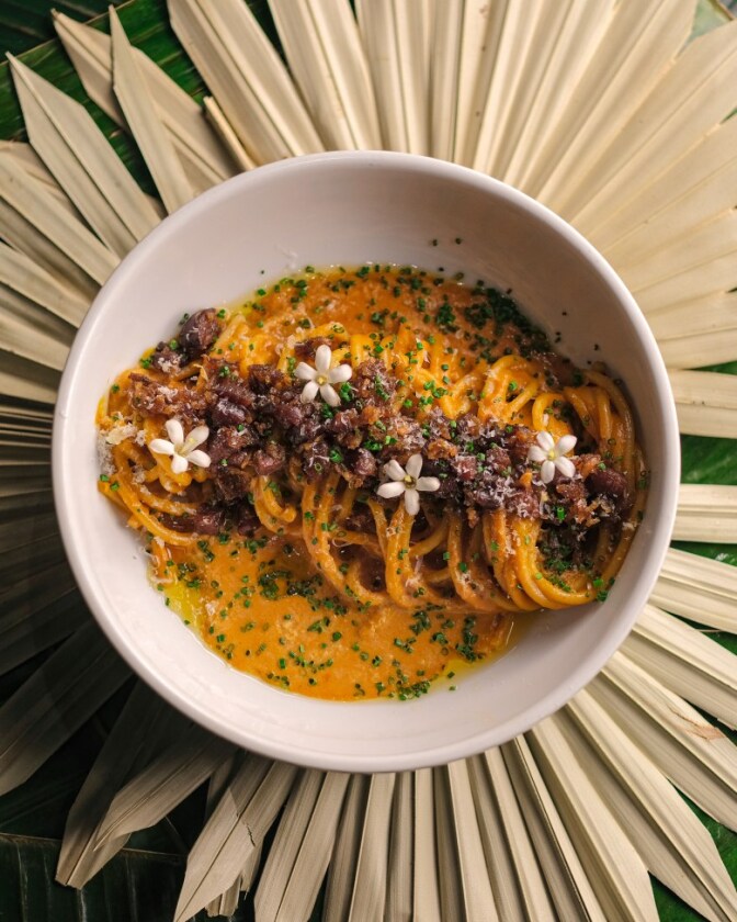 A white bowl containing thick spaghetti noodles tossed in an orange-tinted sauce, topped with crumbled dark red longganisa sausage, chopped chives, and small white jasmine-like flowers.