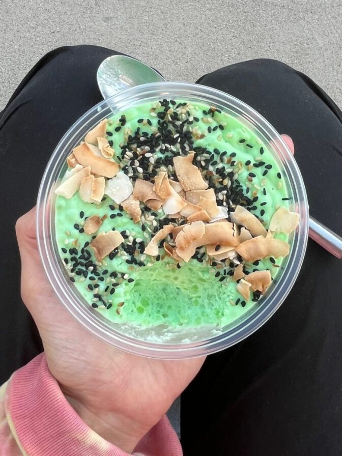 A close up of a person's hands holding a bowl filled with a green dish topped with sesame seeds and other items.