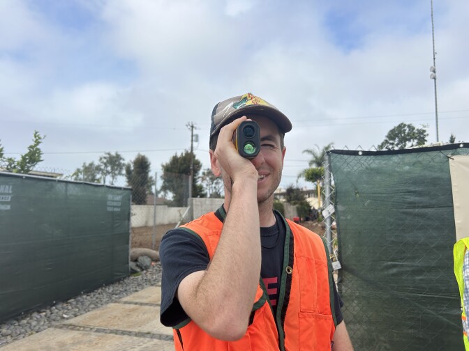 A young man with light skin wearing a neon orange safety vest looks through a monocular. 