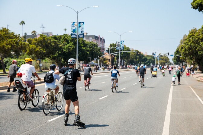 Dozens of bicyclists and rollerskaters travel down an empty, tree-lined road. 