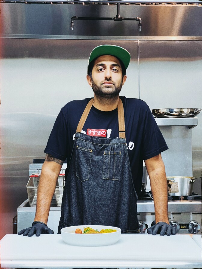 A man with a dark skin tone stands behind a prep counter, wearing a denim apron and a cap, with a bowl of Hokkaido Soup Curry in front of him. His black t-shirt reads "Susukino" in Japanese characters.