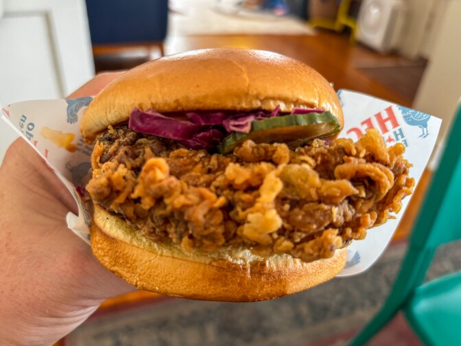 A hand with a light skin tone holds an HFC fried chicken sandwich wrapped in branded paper, showing the full cross-section of the sandwich — a dramatically craggy, amber-colored zangi-style fried chicken cutlet topped with purple cabbage slaw and pickled green cucumber on a golden brioche bun.