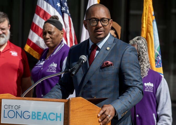 Long Beach Mayor Rex Richardson, a man with dark skin tone, wearing a gray checkered suit, red tie, and glasses, speaks behind a podium with signage that reads "City of Long Beach." There are people standing behind him in front of flags.