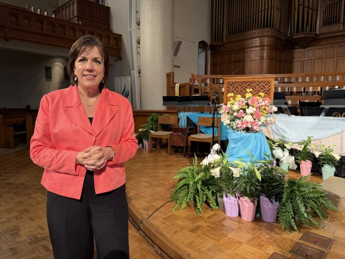 A woman with light skin tone and short brown hair clasps her hands. She is wearing black pants and a salmon pink blazer. 