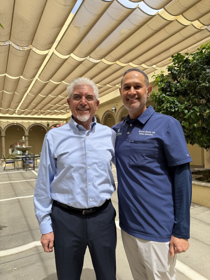 Two men -- one wearing a blue shirt and blue pants and another wearing a blue polo and khaki pants -- interlink arms and look ahead smiling. 