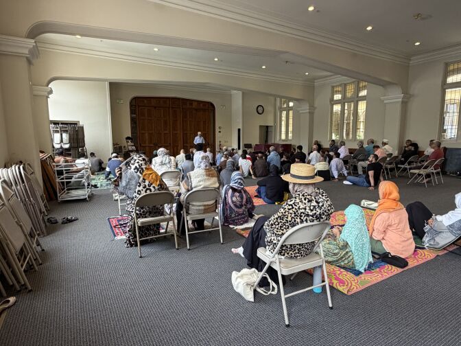 Worshippers sit on rugs and chairs in a large room. 