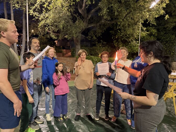 People stand in a circle holding their hands up towards a candle. 