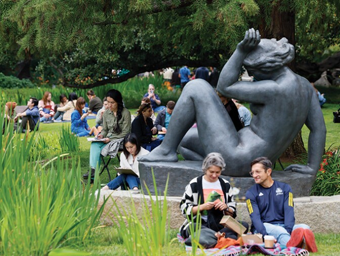 A photograph of several groups of people relaxing on the lawn of the sculpture garden of Norton Simon Museum. The ground is covered with grass and plants with fronds. On the right side photo there is a pine tree, and in front of it is a large gray statute of a nude human figure reclining. People sit around the lawn in different groups talking, eating food, and sketching on paper.