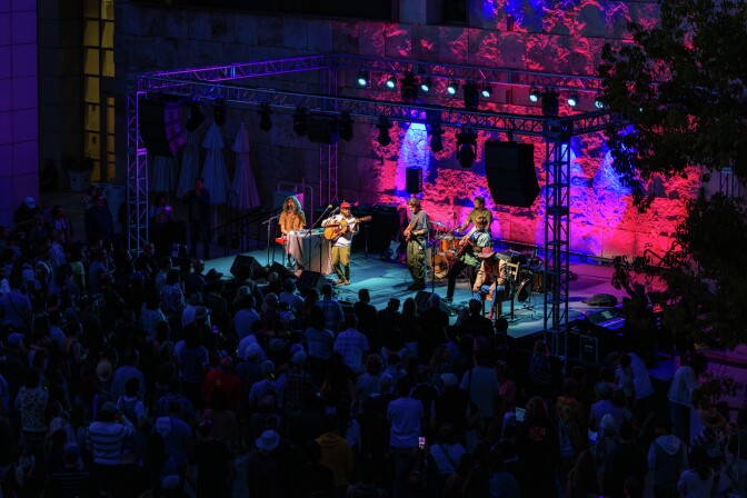 Musicians on an outdoor stage with a metal scaffolding frame around them, a stone wall lit red in the background, and a crowd in darkness, surrounding the stage on three sides.