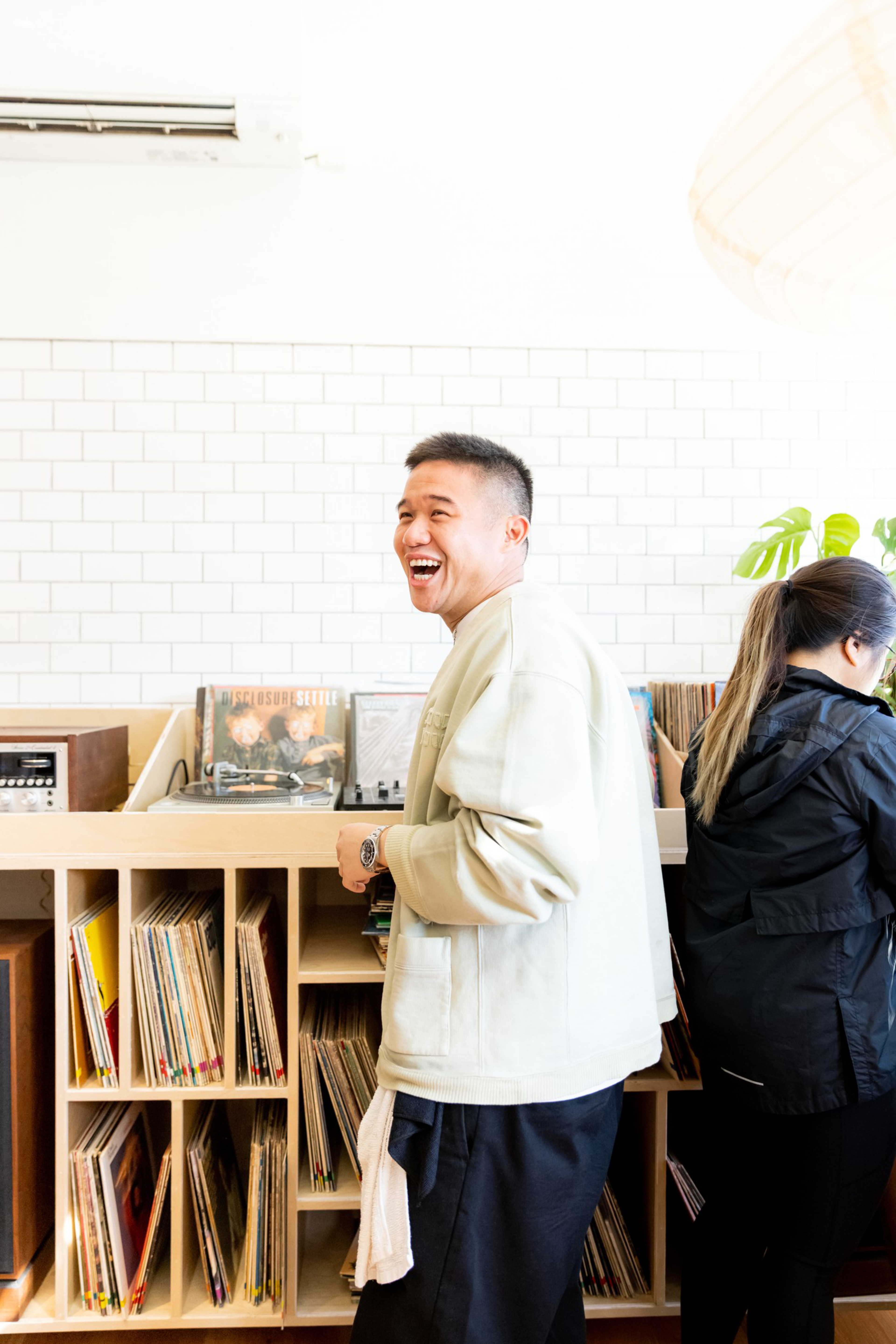 A man in a light jacket laughs while standing near a woman browsing vinyl records in a bright room with shelves full of records.