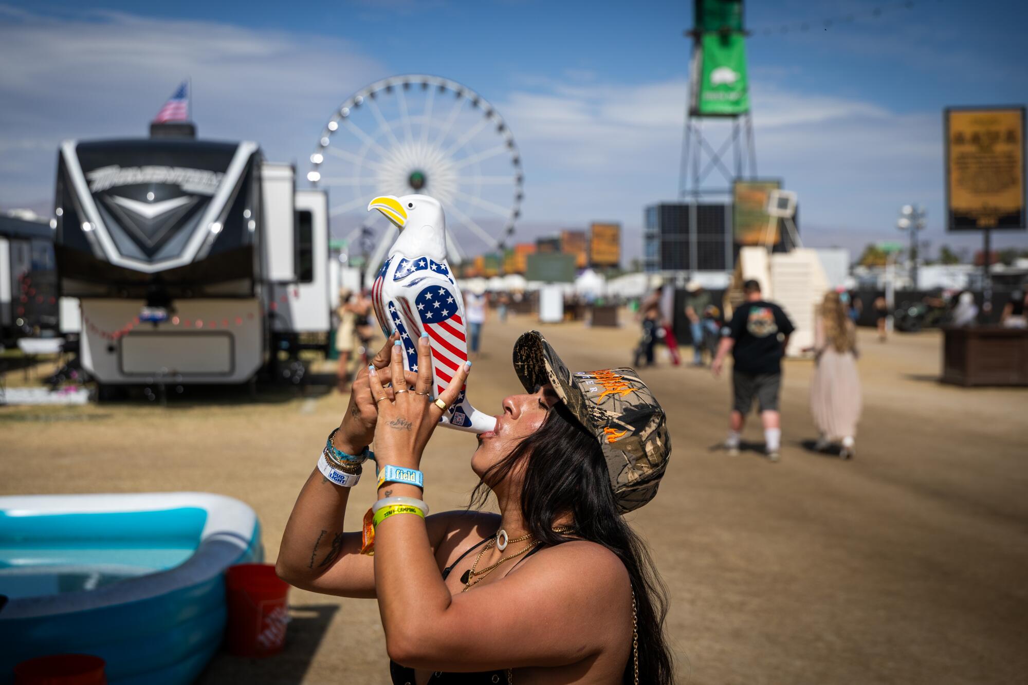 A woman drinks beer out of a beer bong shaped like an eagle with RVs and a Ferris wheel behind her at Stagecoach.