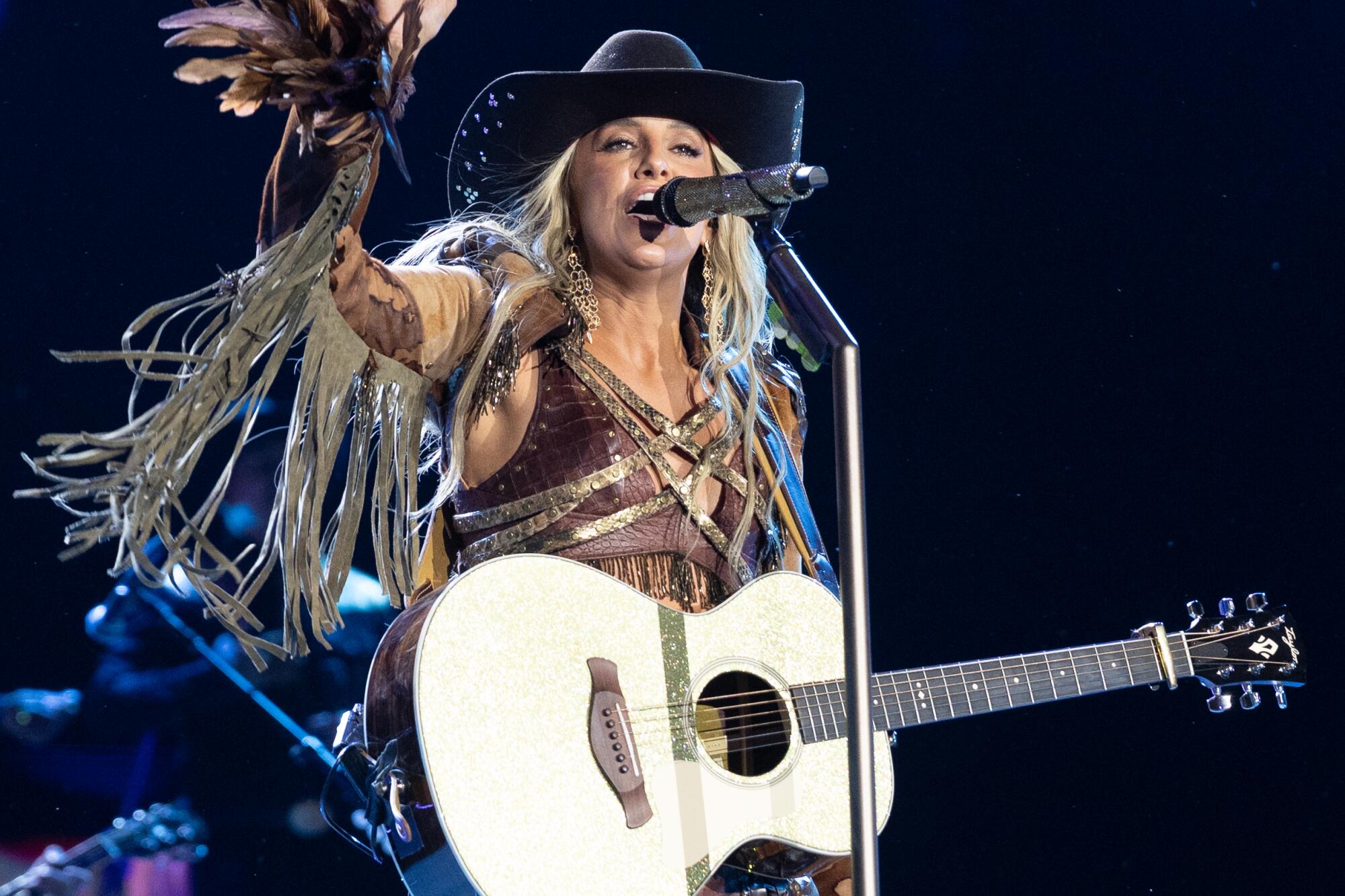 Lainey Wilson wears an outfit with fringe and holds a guitar behind a microphone